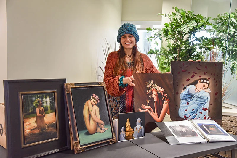 Artist and instructor Ashley Kostak smiling beside a table of her framed oil paintings